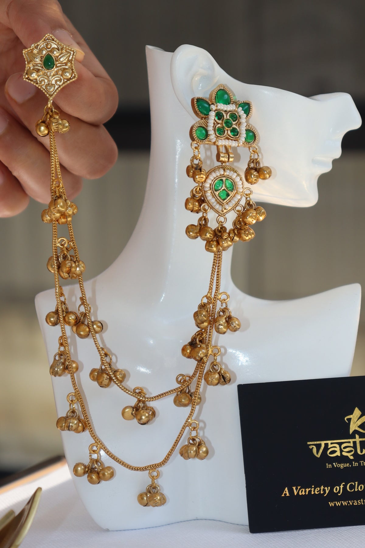 Gold-toned traditional Indian jewelry set with a necklace and matching earrings, featuring green stones and dangling gold bells, displayed on a white mannequin head with a hand holding one earring.
