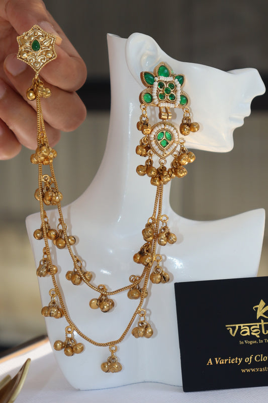 Gold-toned traditional Indian jewelry set with a necklace and matching earrings, featuring green stones and dangling gold bells, displayed on a white mannequin head with a hand holding one earring.