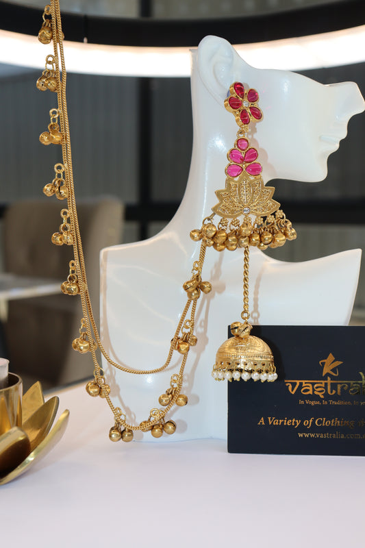 A gold-toned traditional Indian jhumka necklace with red floral stones and a dangling bell, displayed on a white mannequin bust next to a black brand card.