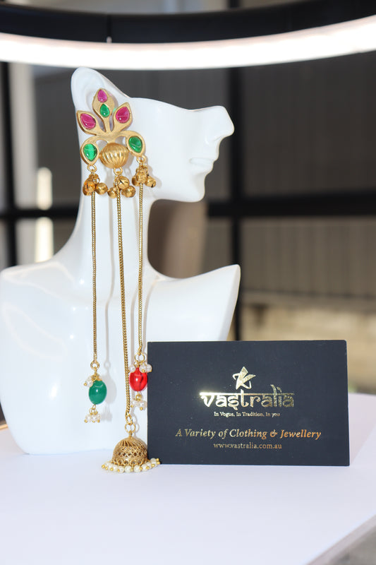 A long gold-toned traditional Indian jhumka earring with green and red stone embellishments, displayed on a white mannequin head next to a black Vastraia business card.