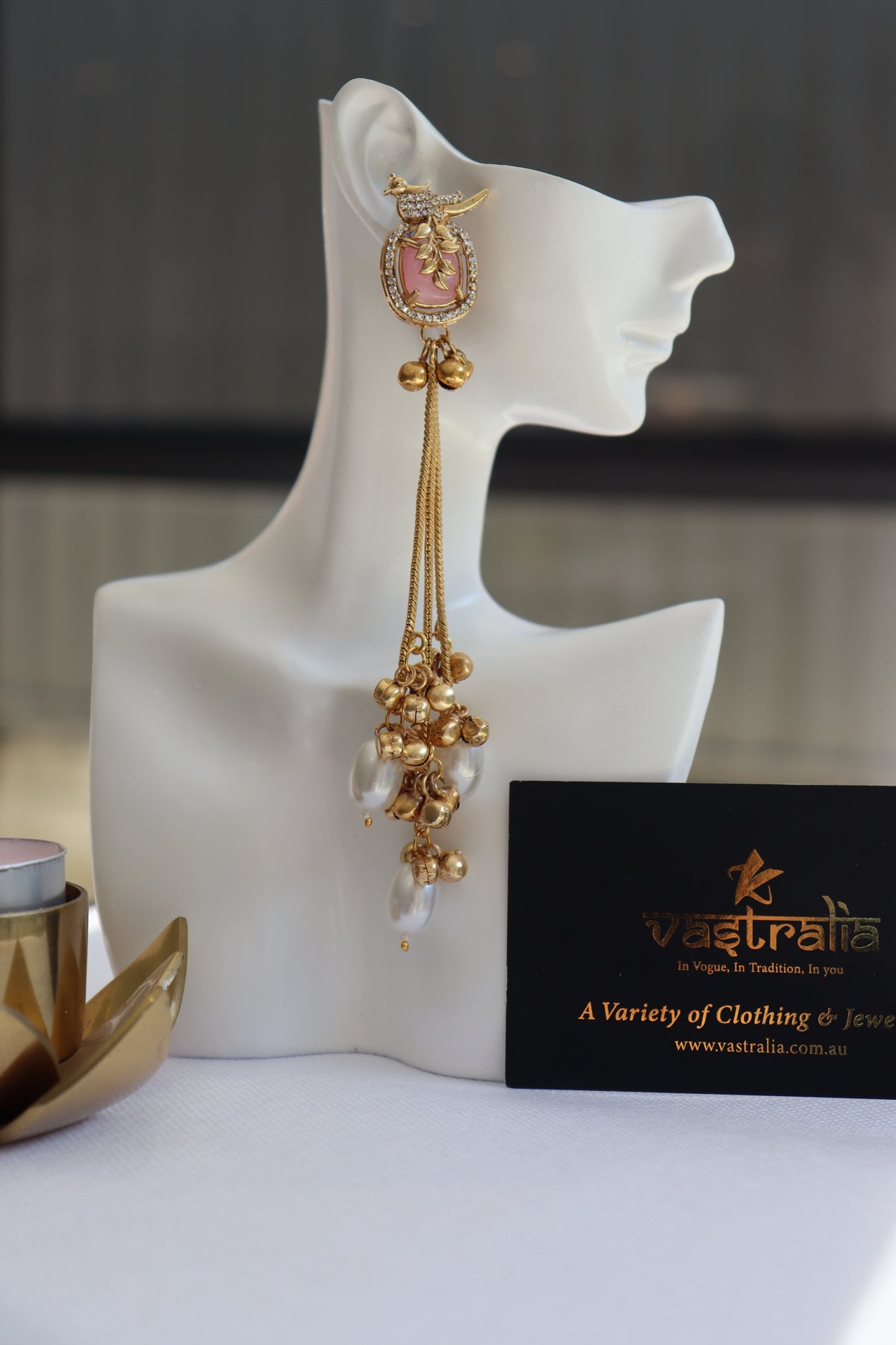 A long gold-toned traditional Indian earring with a pink stone and white pearl drops, displayed on a white mannequin head. A black card with the brand name Vaastraelo is visible next to it.