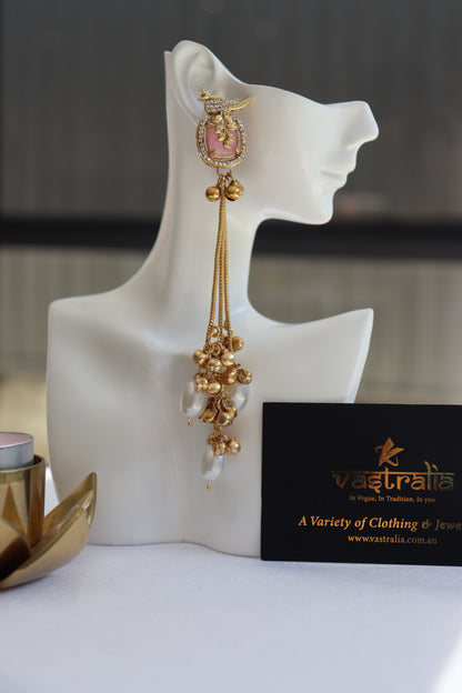 A long gold-toned traditional Indian earring with a pink stone and white pearl drops, displayed on a white mannequin head. A black card with the brand name Vaastraelo is visible next to it.