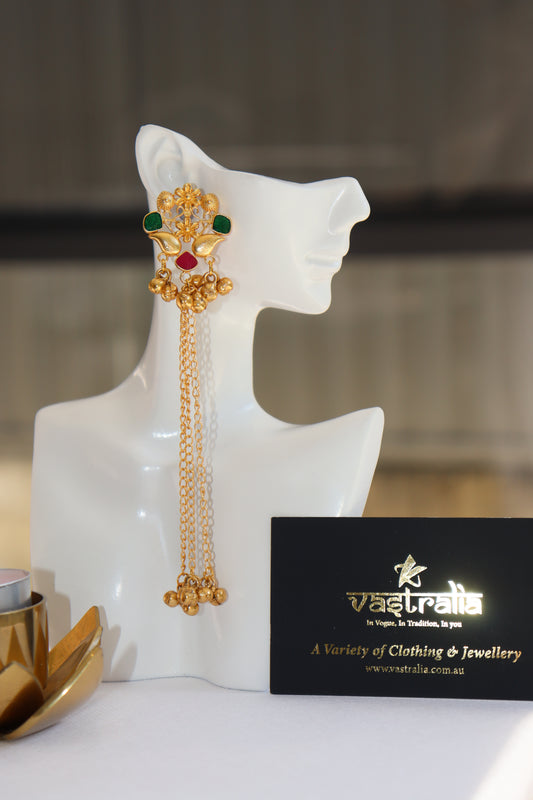 A long gold-toned traditional Indian earring with green and red stones and dangling chains, displayed on a white mannequin bust next to a black branded card.