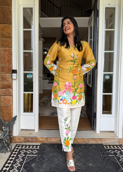 A woman stands in a doorway wearing a mustard yellow embroidered kurta with colorful floral patterns and white pants with matching floral embroidery. The setting is a home entrance with brick walls and glass doors.