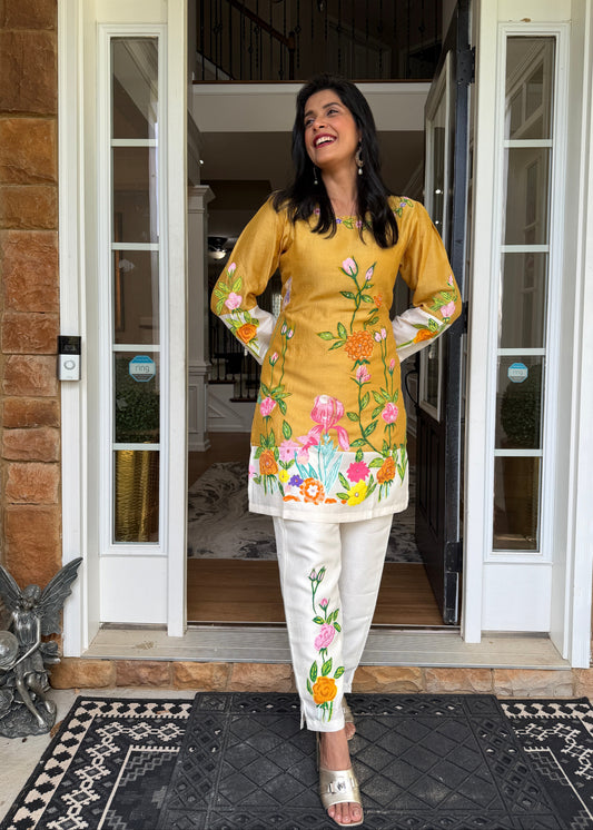 A woman stands in a doorway wearing a mustard yellow embroidered kurta with colorful floral patterns and white pants with matching floral embroidery. The setting is a home entrance with brick walls and glass doors.