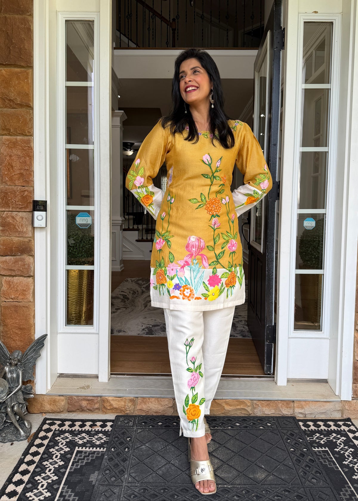 A woman stands in a doorway wearing a mustard yellow embroidered kurta with a white border and colorful floral print, paired with white pants featuring similar floral embroidery. The setting is a home entrance with brick walls and a black patterned rug.