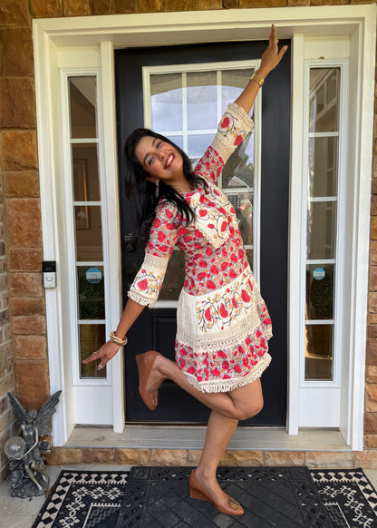 A woman posing in a red and white floral printed dress with cream lace trim, standing on a front porch in front of a black door.