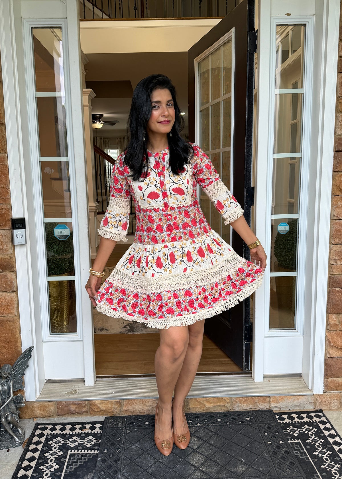 A woman stands in a doorway wearing a short, flared embroidered dress with three-quarter sleeves. The dress has a cream base with red and pink floral embroidery and white lace trim.
