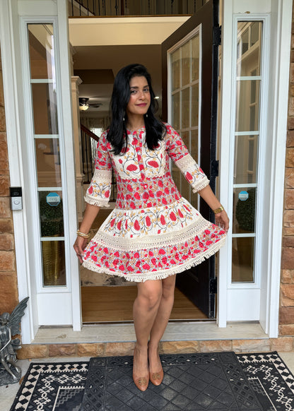 A woman stands in a doorway wearing a short, flared embroidered dress with three-quarter sleeves. The dress has a cream base with red and pink floral embroidery and white lace trim.