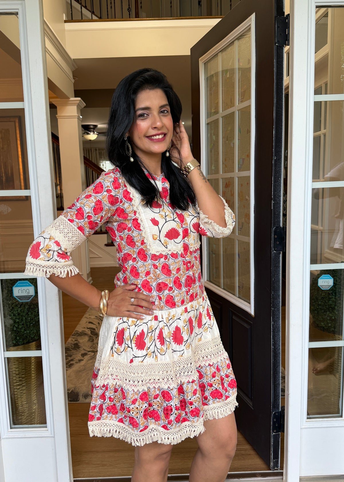 A woman modeling a short, red and cream floral printed dress with lace trim, standing in an open doorway of a building.