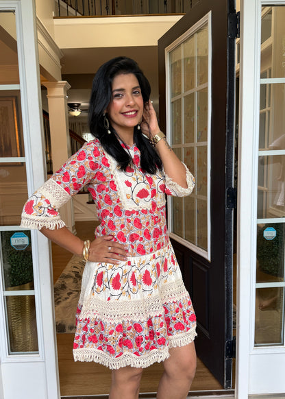 A woman modeling a short, red and cream floral printed dress with lace trim, standing in an open doorway of a building.