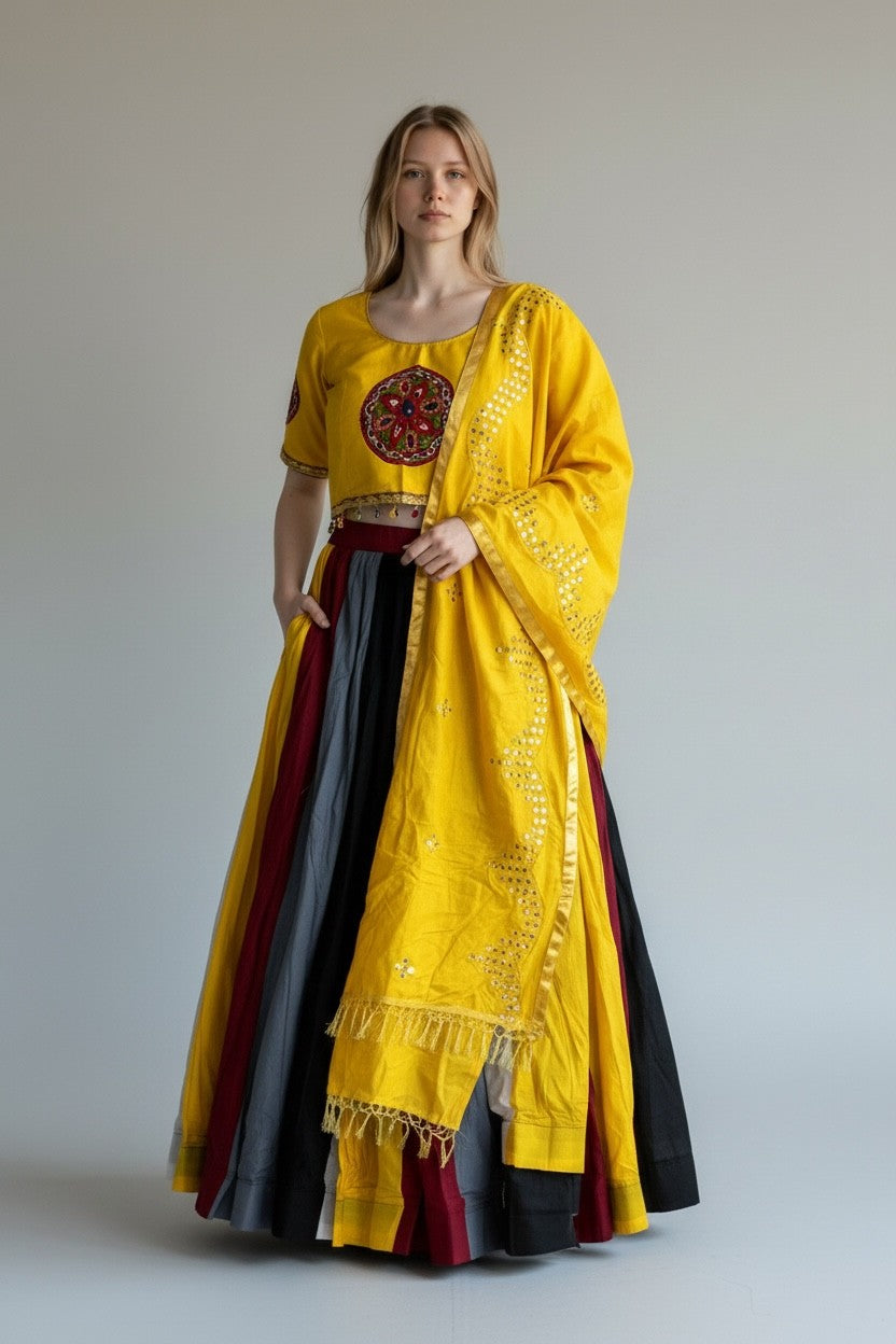 A woman modeling a traditional Indian outfit consisting of a bright yellow kurta with a red and gold circular embroidered motif on the chest, paired with a matching yellow dupatta and a layered skirt in black, gray, and maroon. The model is standing against a plain light gray studio background.