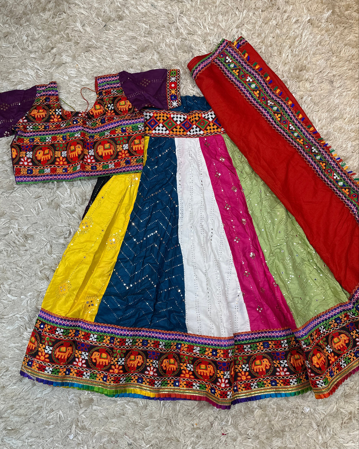 A traditional Indian ethnic outfit laid flat on a beige shag rug, featuring a multicolor skirt with yellow, blue, white, pink, and green panels, a purple embroidered blouse, and a red dupatta with colorful borders.