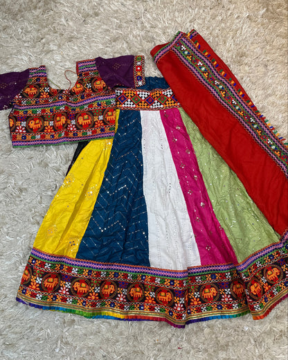 A traditional Indian ethnic outfit laid flat on a beige shag rug, featuring a multicolor skirt with yellow, blue, white, pink, and green panels, a purple embroidered blouse, and a red dupatta with colorful borders.