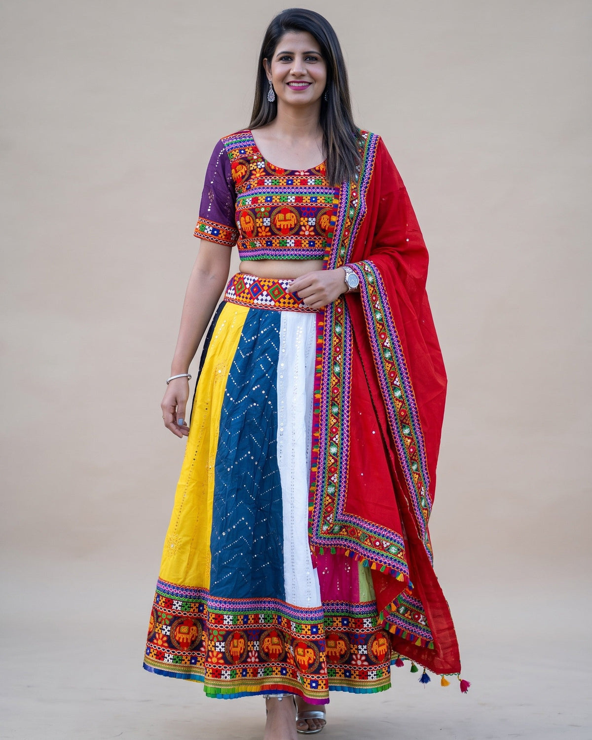 A woman modeling a vibrant, multicolored traditional Indian ethnic outfit consisting of a heavily embroidered purple blouse, a multi-colored skirt with yellow, blue, and white panels, and a bright red dupatta with gold borders, set against a plain beige studio background.