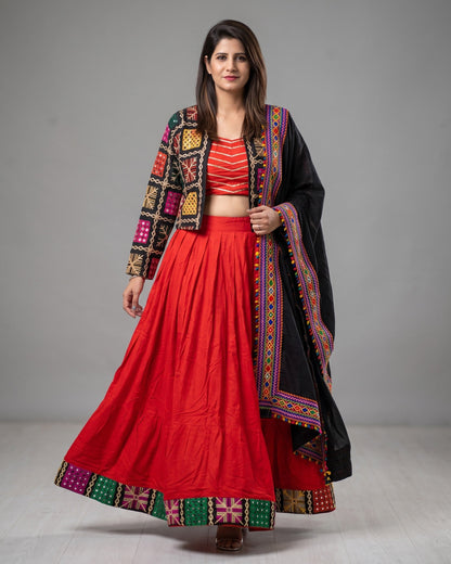 A woman modeling a traditional Indian outfit consisting of a bright red flared skirt (lehenga), a red and white striped crop top, a black embroidered jacket, and a black dupatta with colorful borders, set against a plain gray studio background.