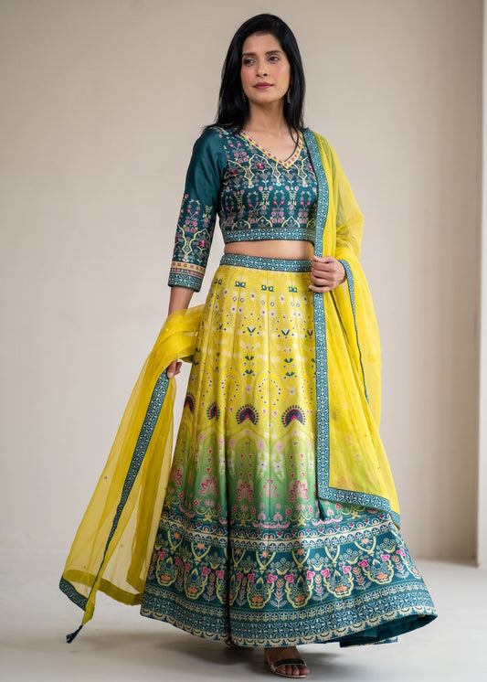 A woman modeling a traditional Indian lehenga choli set with a yellow skirt, teal embroidered blouse, and a yellow dupatta with teal borders, set against a plain beige studio background.