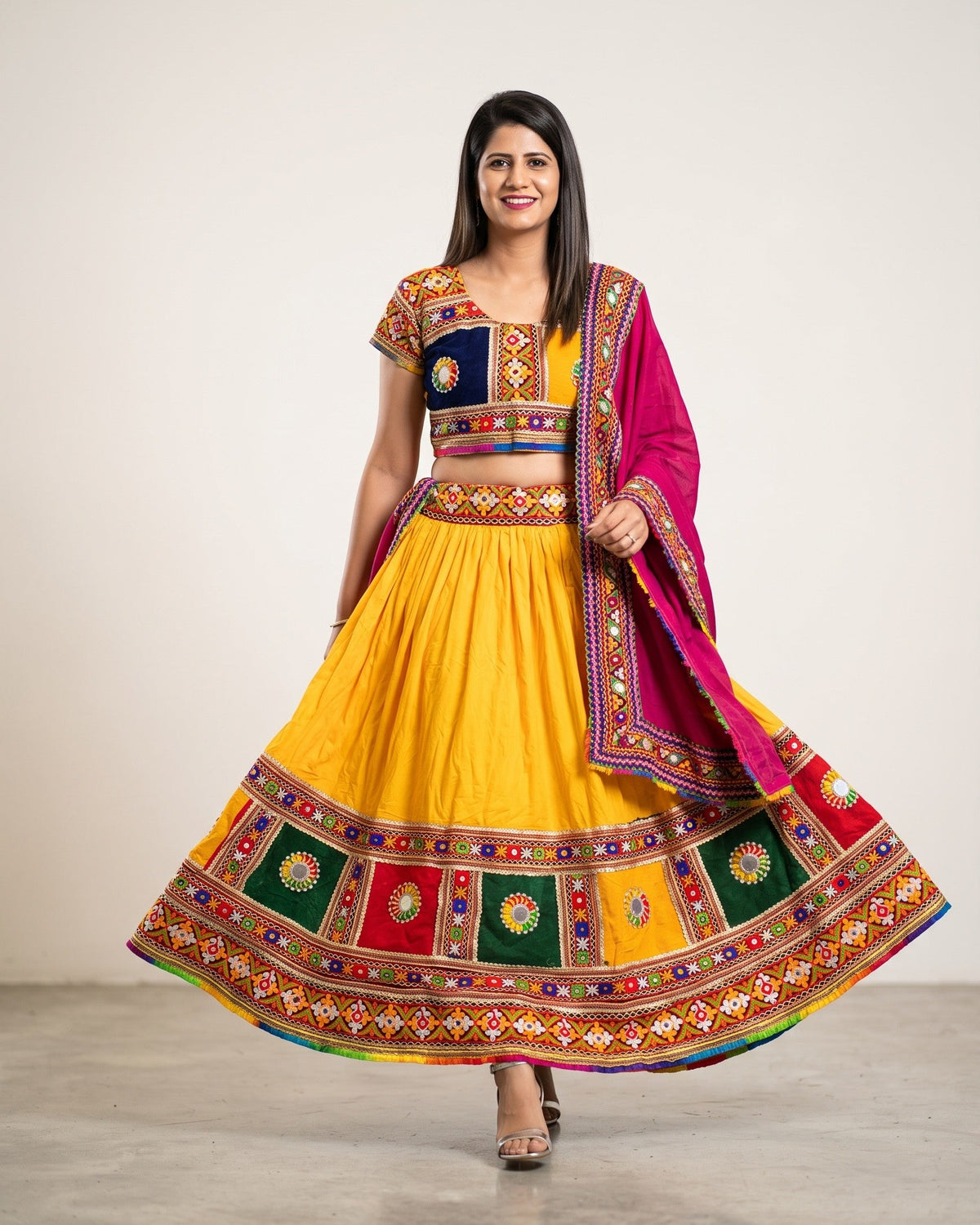 A woman modeling a traditional Indian anarkali gown with a bright yellow skirt, a navy blue embroidered bodice, and a magenta dupatta with colorful geometric borders, set against a plain studio background.