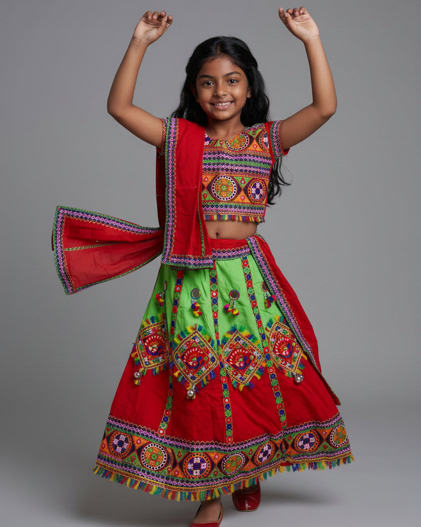 A young girl modeling a traditional Indian ethnic outfit consisting of a bright red and lime green skirted dress with colorful embroidered patterns and a matching red dupatta, shot against a solid gray studio background.