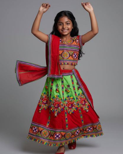 A young girl modeling a traditional Indian ethnic outfit consisting of a bright red and lime green skirted dress with colorful embroidered patterns and a matching red dupatta, shot against a solid gray studio background.