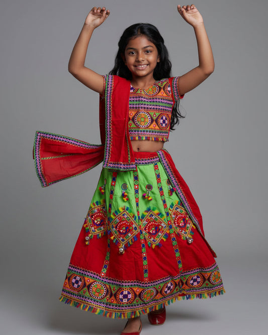 A young girl modeling a traditional Indian ethnic outfit consisting of a bright red and lime green skirted dress with colorful embroidered patterns and a matching red dupatta, shot against a solid gray studio background.