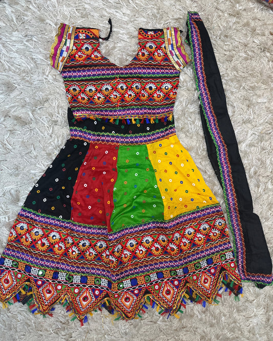 A colorful embroidered traditional dress laid flat on a white shag rug, with a matching black dupatta draped alongside it. The dress features a multicolor patterned bodice, three vertical color-blocked panels in black, red, and green, a bright yellow panel, and extensive mirror work and embroidery along the hem and sleeves.