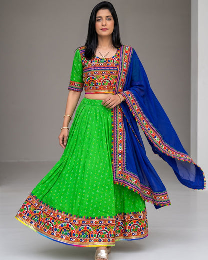 A woman modeling a traditional Indian ethnic wear set consisting of a bright green flared skirt with a heavily embroidered multicolor border, a matching green short-sleeve blouse with a colorful embroidered yoke, and a royal blue dupatta draped over her shoulder.
