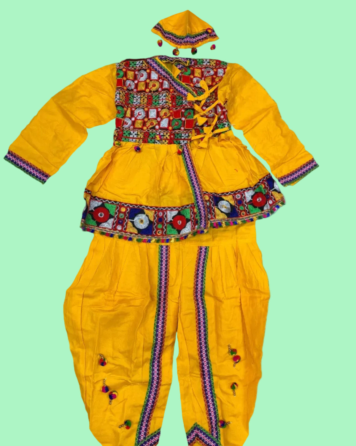A bright yellow traditional outfit set laid flat on a white shag rug, featuring a long-sleeve embroidered top with a colorful patchwork-style chest panel, decorative waist trim, matching pants with vertical embroidered trim, and a matching cap. A packaged similar item and a small accessory are partially visible at the edges.
