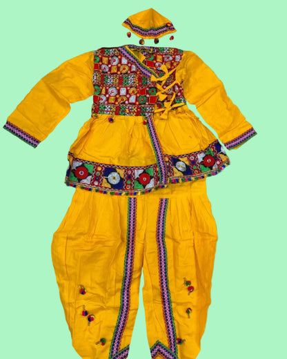 A bright yellow traditional outfit set laid flat on a white shag rug, featuring a long-sleeve embroidered top with a colorful patchwork-style chest panel, decorative waist trim, matching pants with vertical embroidered trim, and a matching cap. A packaged similar item and a small accessory are partially visible at the edges.