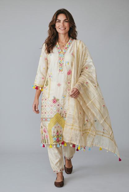 A woman modeling a traditional three-piece salwar kameez suit set in cream, featuring colorful floral embroidery, yellow geometric patterns, and multicolored tassels, paired with a matching sheer dupatta.
